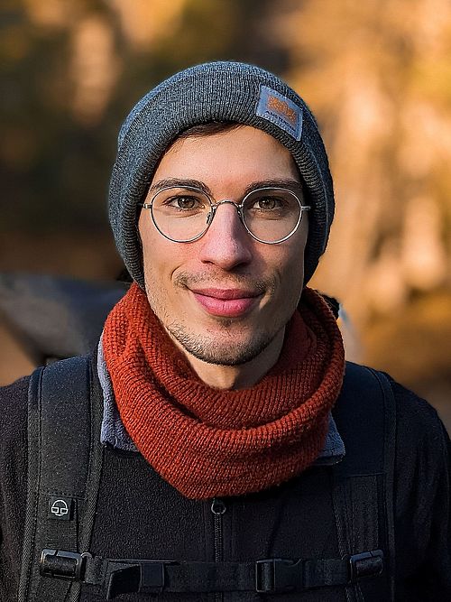 A potrait image of Aramis Keller wearing a black coat, a backpack, a red scarf and a hat. He wears glasses. Background is blurred. Image is taken from the front.